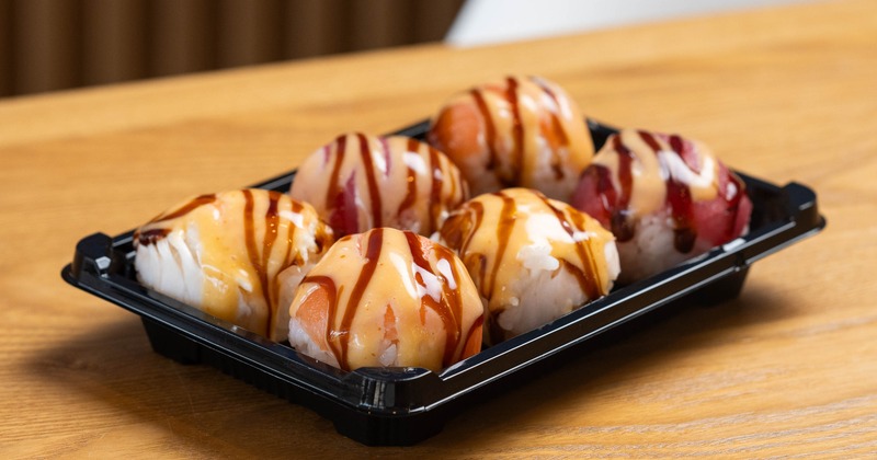 Black plastic takeout container filled with six round sushi balls topped with sauces