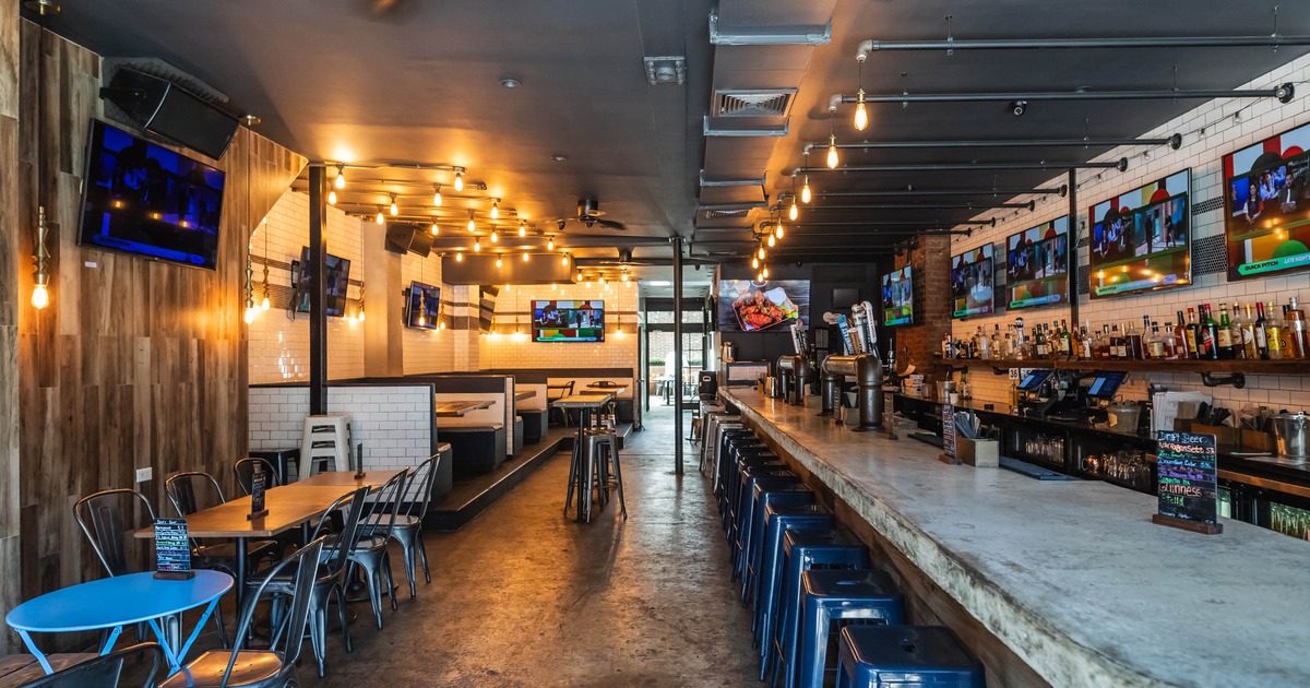 Interior of a modern bar and restaurant featuring a long stone bar, metal stools, and cozy booths with televisions