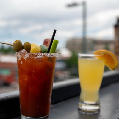 Two cocktails sit on a rooftop ledge with a blurred city tower in the background.