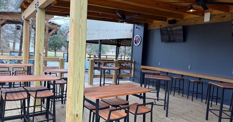 Covered outdoor patio with wooden tables, tall stools, and ceiling fans