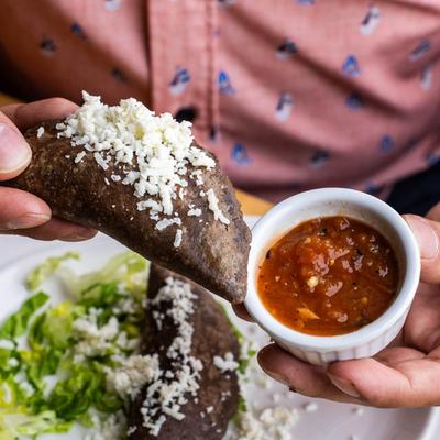A hand dipping a cheese-covered blue-corn empanada into red salsa.