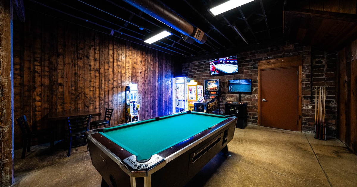 Game room with a green pool table in the center, arcade machines along the wooden walls, seating