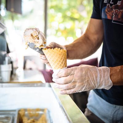 An employee standing  by parlor with a scoop of ice cream in one hand and a cone in the other.