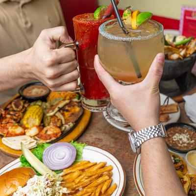 Two hands toasting with colorful cocktails over a table with various dishes.