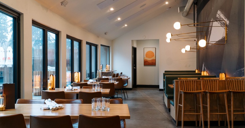 Modern restaurant interior with large windows, wooden tables, leather chairs, and green booths