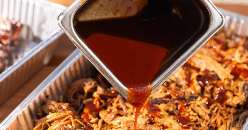 Rich, dark barbecue sauce being poured from a metal container onto a tray of shredded pulled pork