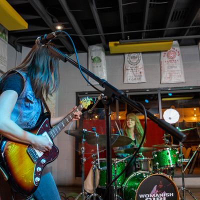 Band on stage with female playing guitar in foreground and female drumming in the background.