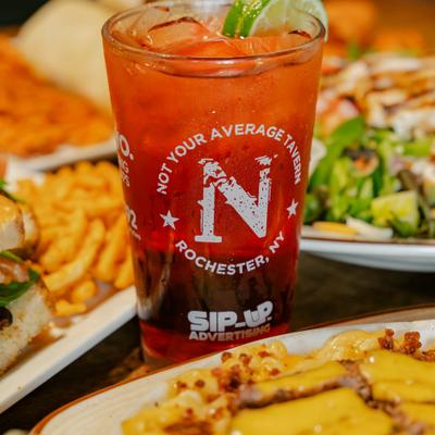 Red cocktail in a branded pint glass, served alongside various dishes.