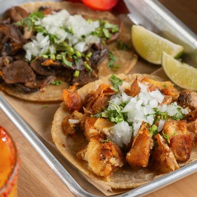Street tacos served in a metal tray with diced onion, cilantro and lime wedges.