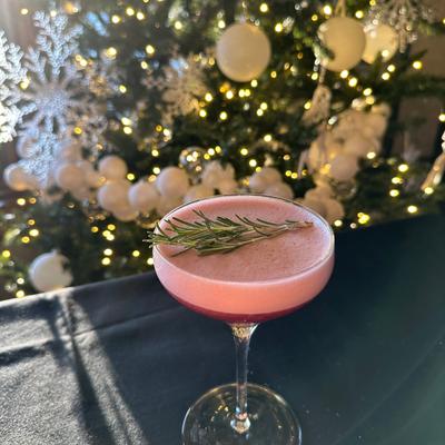 Pink cocktail with a rosemary garnish on a black table, with warmly lit Christmas tree.
