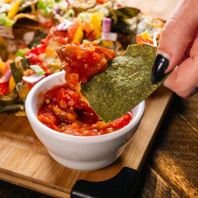 A hand dipping a green tortilla chip into red salsa.