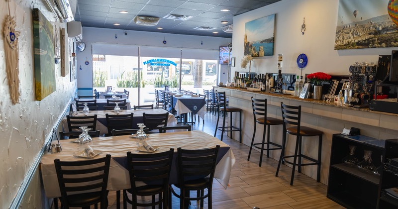 Interior of a restaurant featuring tables with white tablecloths, a bar counter with bottles