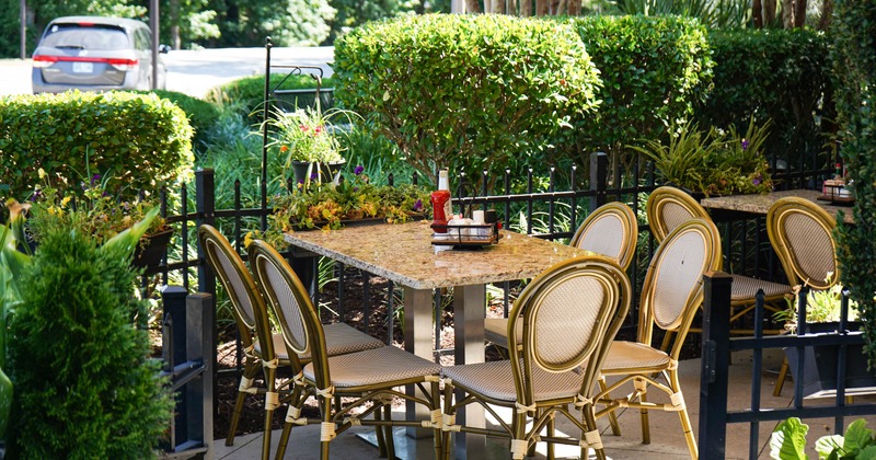 An outdoor setting - a granite table & six gold metal chairs, surrounded by lush greenery.
