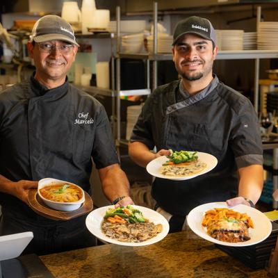 Two professional chefs holding plated dishes in a kitchen setting.
