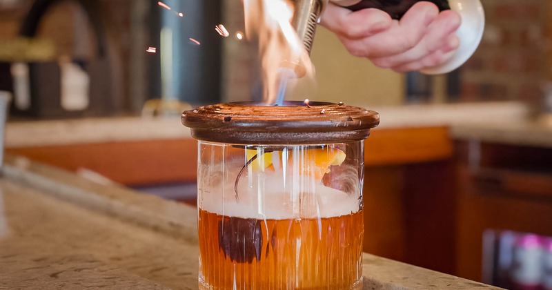 A bartender using a bar torch to prepare Smoked Old Fashioned