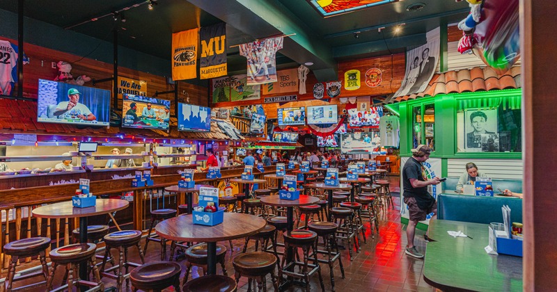 Interior of a sports-themed bar with tables, stools, banners, and multiple TVs
