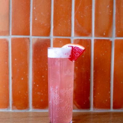 Tall glass of pink Paloma, with a grapefruit wedge, set against a tiled orange backdrop.