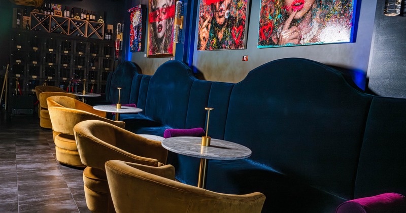 Lounge interior with velvet seating, marble tables, colorful paintings, and a wine rack