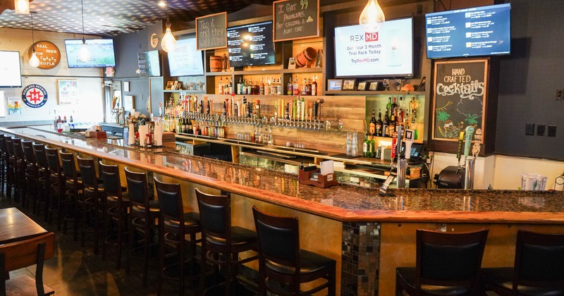 A spacious bar interior, long granite countertop, wooden stools, shelves with liquor bottles