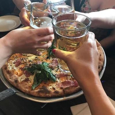 Hands clinking glasses in a group toast above a pizza on a table.