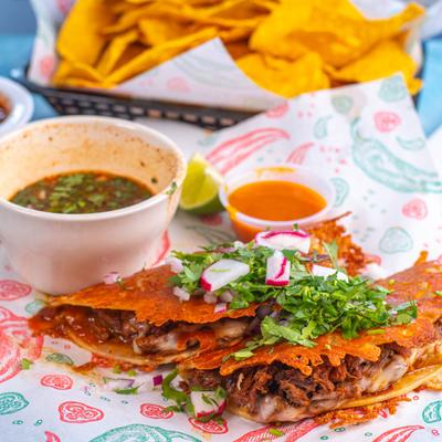 Birria tacos served with dipping broth, salsa, and chips in the background.