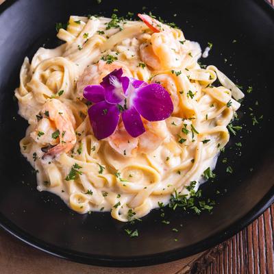 A black bowl of creamy fettuccine topped with shrimp and garnished with a vibrant purple orchid.