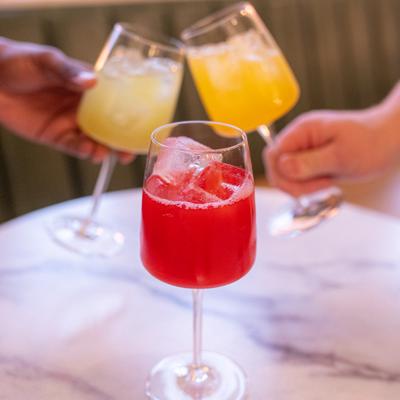 A red aqua fresca sits on a marble table, two hands toast with yellow drinks in the background.