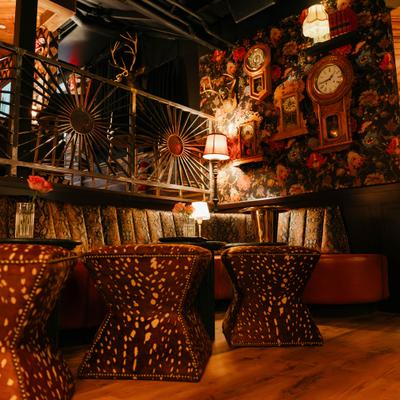 Cozy bar interior with warm lighting, floral wallpaper, vintage clocks, and antlers.