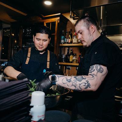 Two chefs mixing a salad in a large metal bowl.