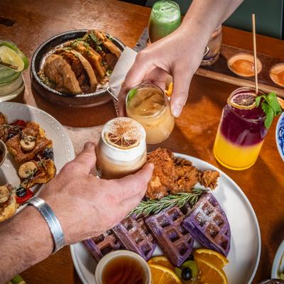 Hands clinking drinks over a table of colorful brunch dishes.