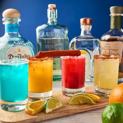 Colorful margaritas on a wooden board, surrounded by tequila bottles, garnished with citrus slices.