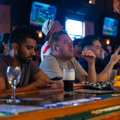 Patrons sit at a crowded bar watching sports with drinks nearby.