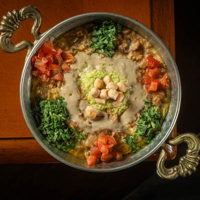 Ful medames, a dish of cooked fava beans served in a metal bowl with ornate handles.