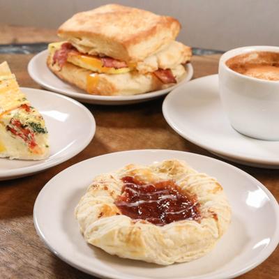 Strawberry danish, a slice of quiche, a breakfast sandwich on a biscuit, and a cup of espresso.