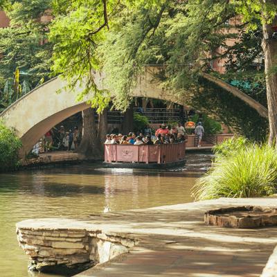 Riverside dining patio beneath lush green trees and a stone bridge.