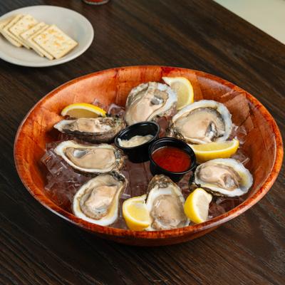 Raw oyster platter served on ice in a wooden bowl with lemon wedges and dipping sauces.