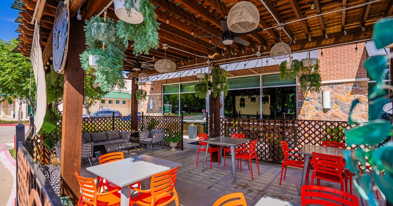 Outdoor restaurant patio with red-orange chairs, gray tables, hanging plants, decorative lighting