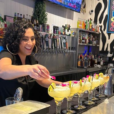 picture of smiling female bartender garnishing 6 margaritas on a bar.