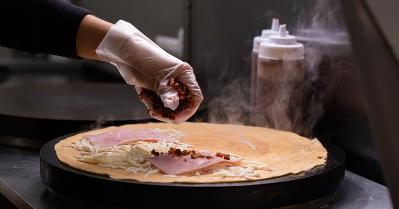A hand topping a savory crepe with bacon bits