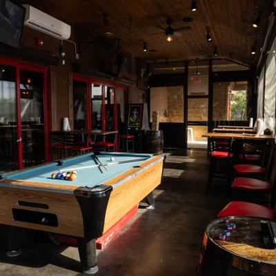 Interior with a pool table lit by warm sunlight, tables and red chairs.