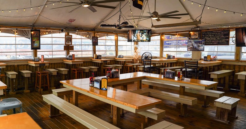 Indoor dining area with wooden tables and benches, large windows, and mounted televisions