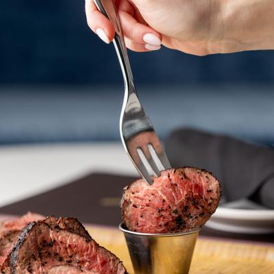 A hand using a fork to dip a slice of medium-rare steak into a cup pf sauce.
