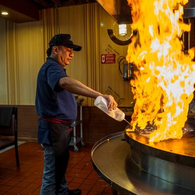 A chef squirts liquid onto a grill, igniting a massive, roaring flame.