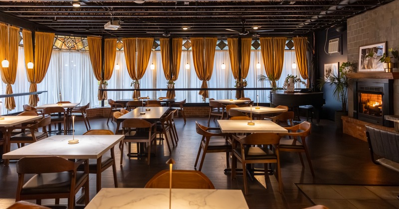 A warmly lit interior with tables, lit candles, large windows with gold curtains and a lit fireplace