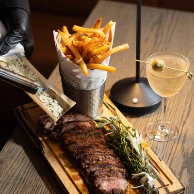 Bone marrow being topped over the steak served with fries and a cocktail.