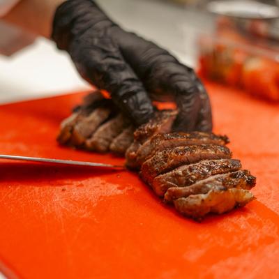 A hand in a black glove slicing a steak on the red cutting board.