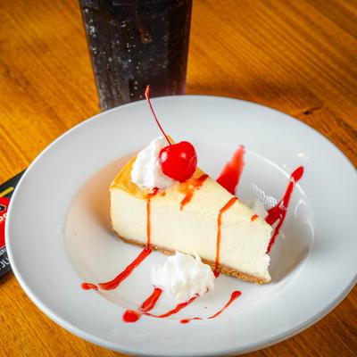 A slice of cheesecake, garnished with whipped cream and a cherry, and drizzled with a red sauce.