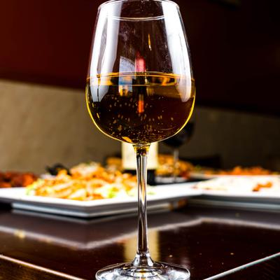 A glass or white wine on a dark table with food plates in the background.
