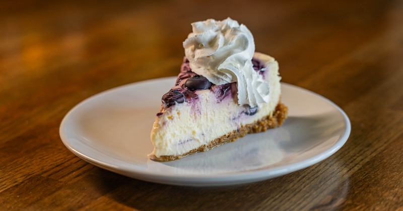 A slice of blueberry lemon cheesecake sits on a white plate atop a wooden table