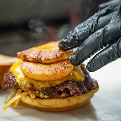 Gourmet smash burger being topped with grilled pineapple, close-up.